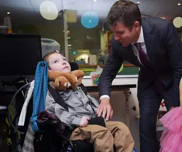 A man in a suit leans over to interact with a child in a wheelchair, with colorful balloons in the background.