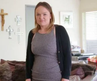 A woman in a striped dress and black cardigan stands in a living room with decorative crosses on the wall.