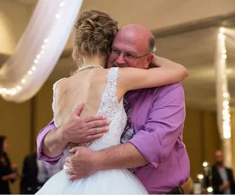 Bride in white dress hugs older man in a purple shirt during a wedding dance, with soft lights in the background.