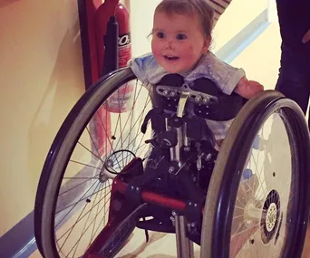 A smiling toddler in a wheelchair, who lost limbs to meningitis, is depicted navigating a hallway.