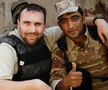 Two men in military gear smiling, one giving a thumbs up, in a desert setting.