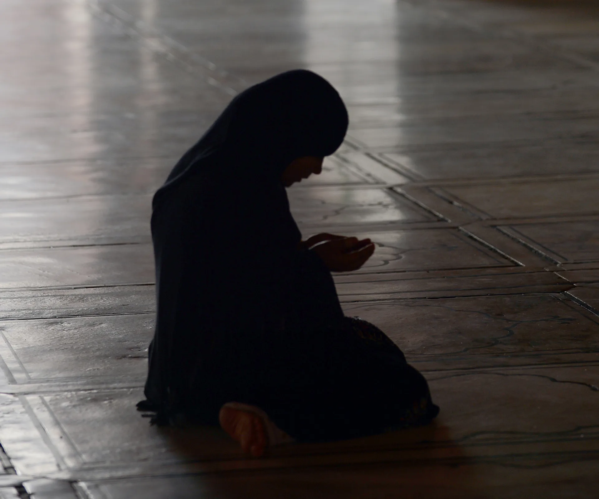 A silhouetted person in a headscarf kneels in prayer on a tiled floor.