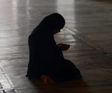 A silhouetted person in a headscarf kneels in prayer on a tiled floor.
