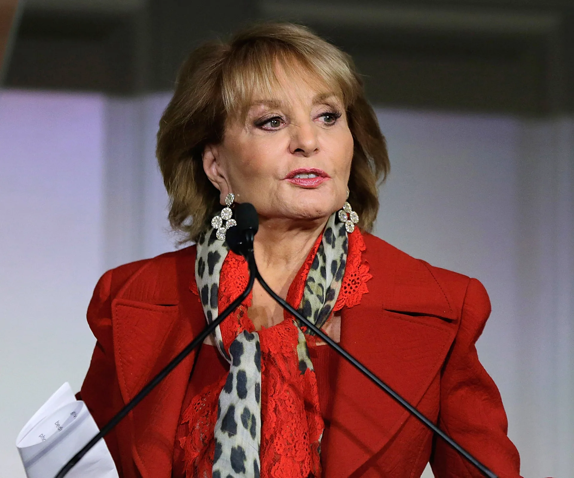 Barbara Walters speaking at an event, wearing a red outfit with a leopard print scarf.
