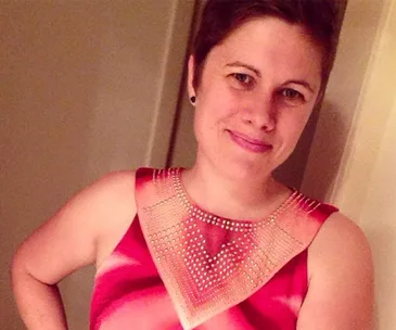 A person in a red and pink dress stands smiling, with a unique design on the top resembling a triangular pattern.