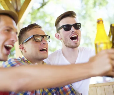 Group of friends wearing sunglasses, cheering with beer bottles outdoors.