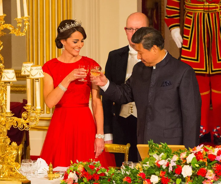 A woman in a red gown and tiara toasts with a man in a formal setting adorned with flowers and candles.
