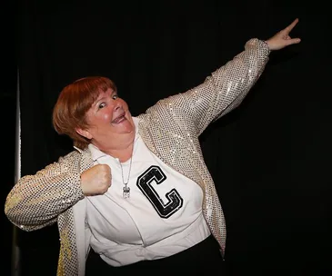 Person posing humorously in a sequined jacket and "C" shirt, mimicking a superhero's stance against a dark background.