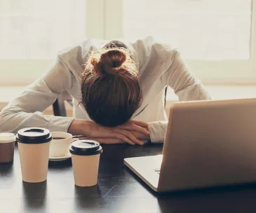 Person resting head on desk beside laptop and coffee, looking stressed.