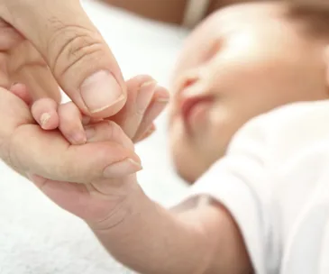 Adult hand holds a baby's tiny hand, with the baby lying down and blurred in the background.