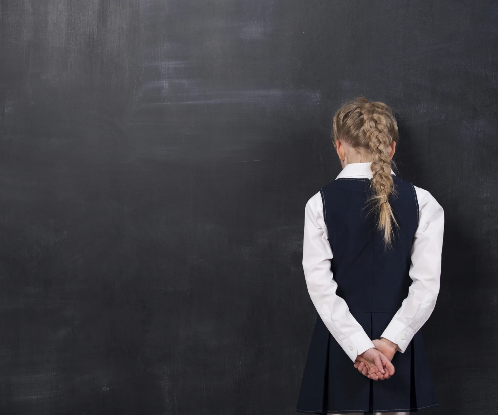 A young girl stands facing a blackboard, hands clasped behind her back, with a long braid.
