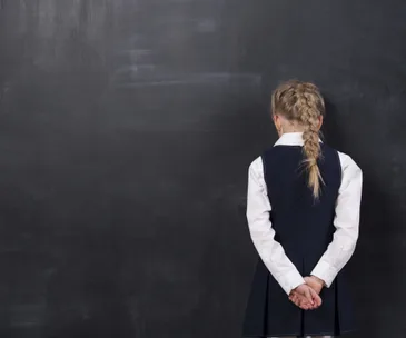A young girl stands facing a blackboard, hands clasped behind her back, with a long braid.