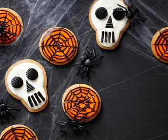 Halloween cookies with skull and spider web designs, decorated with black licorice and plastic spiders on cobweb backdrop.