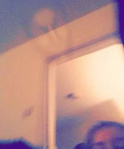 Blurry selfie showing a partially visible face with a ghostly figure appearing above an open doorway.