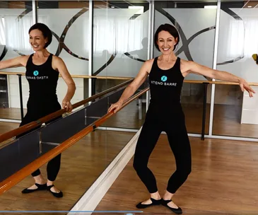 Instructor in a dance studio performing barre exercises, wearing Xtend Barre attire, smiling and holding a barre.