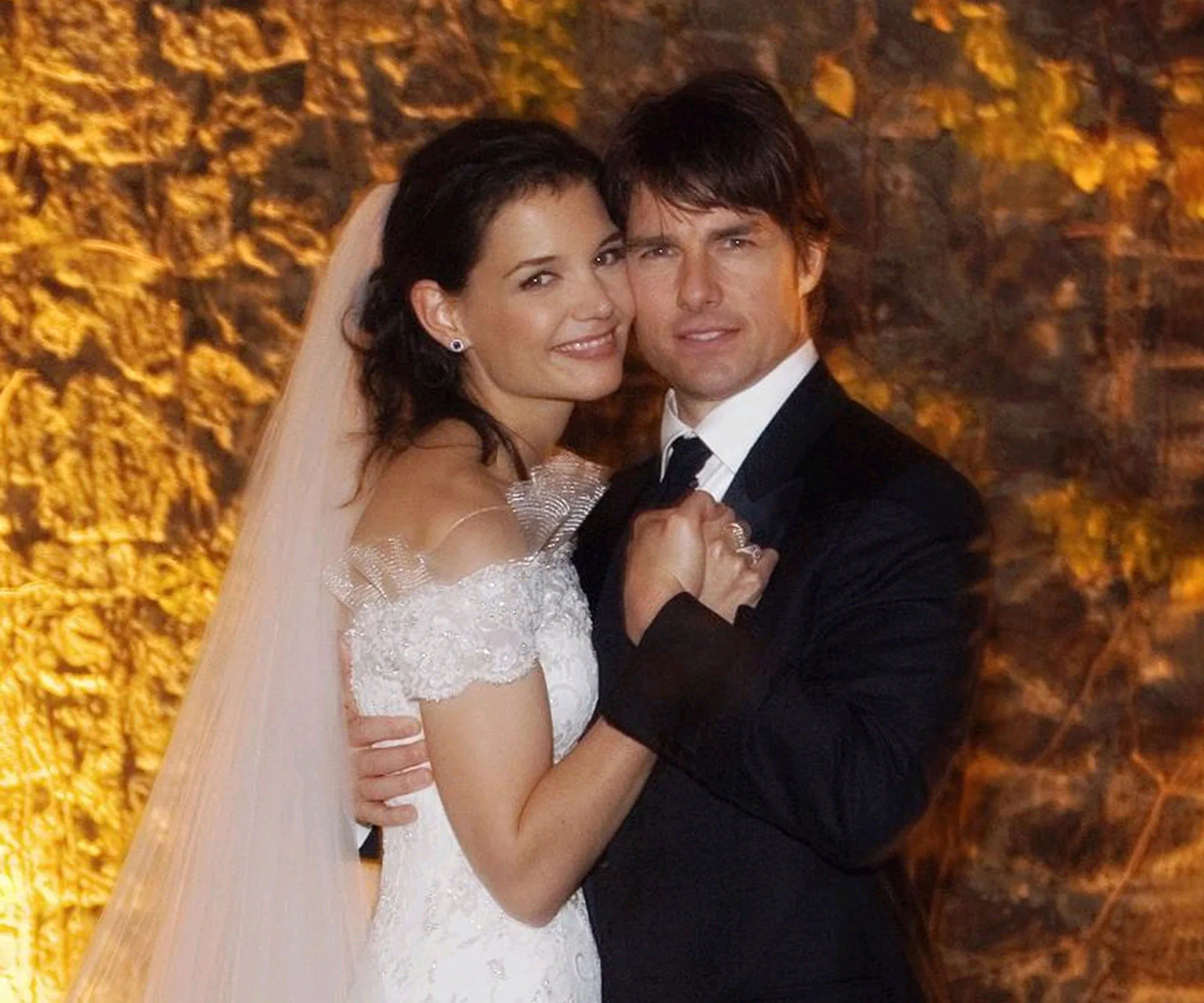 A bride and groom embracing, smiling in a warmly lit setting with a stone backdrop.