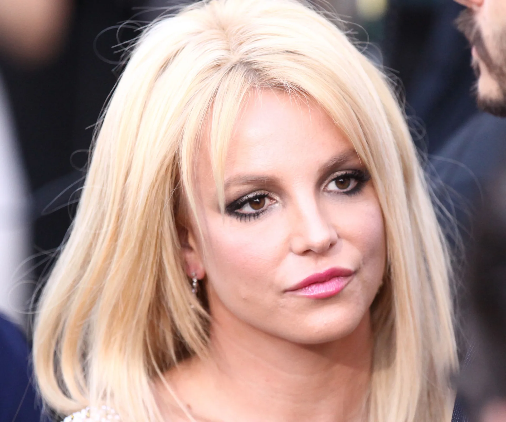 A woman with long blonde hair and a serious expression looks off to the side, surrounded by blurred background figures.