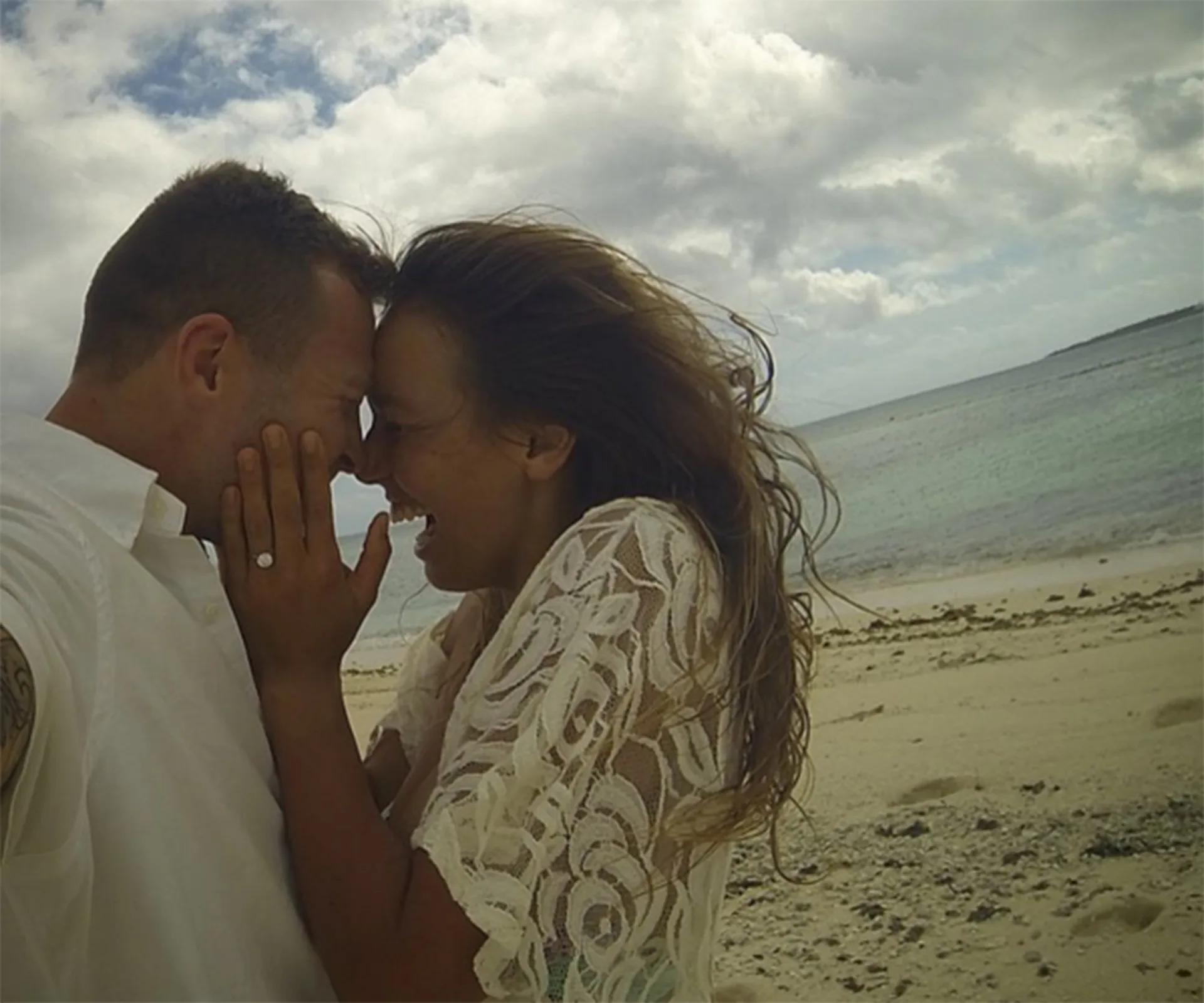 Couple smiling closely on a beach, with a visible engagement ring on the woman's hand.
