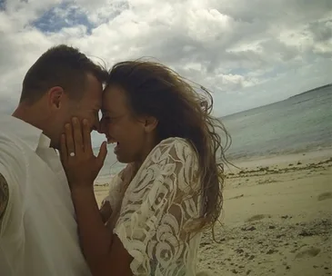 Couple smiling closely on a beach, with a visible engagement ring on the woman's hand.