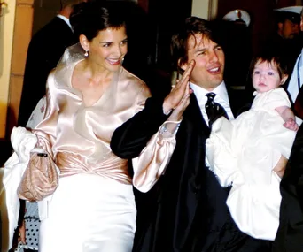 A couple dressed formally smiles as they hold a baby in their arms, during a public event.