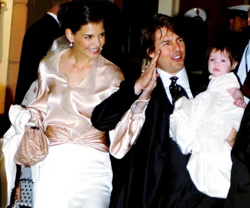 A couple dressed formally smiles as they hold a baby in their arms, during a public event.
