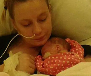 A mother in a hospital bed with a nasal tube holds her newborn baby girl, who is wearing a pink polka dot outfit.
