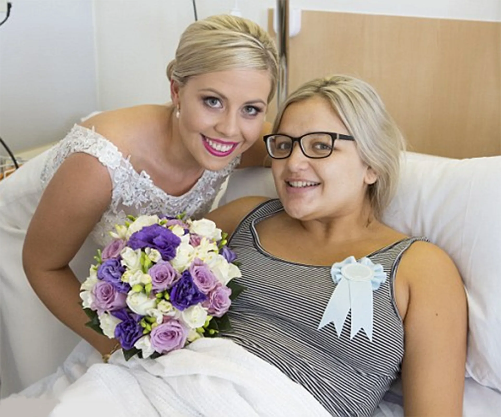 Bride in wedding dress visits a smiling bridesmaid in hospital bed, holding a bouquet.