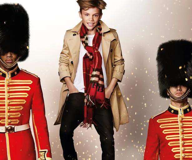 Young model in a Burberry trench coat and scarf stands between two guards in red uniforms and bearskin hats.