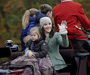 Woman in a green coat and hat waves while holding a child, seated on a carriage with others in outdoor setting.