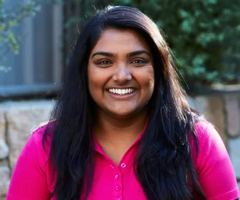 Smiling woman with long dark hair in a pink shirt, outdoors with a blurred background.