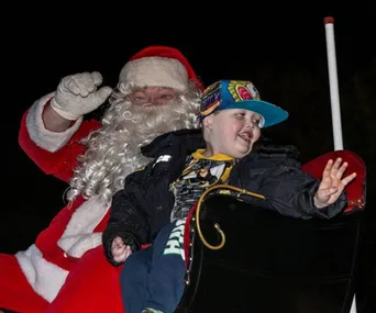 Santa Claus waving with a young boy wearing a colorful cap in a sleigh during an early Christmas celebration at night.