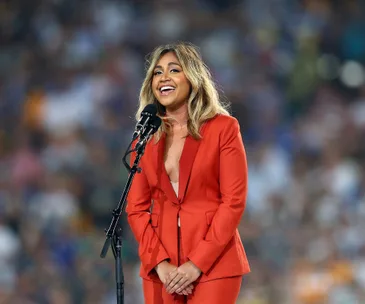 Singer in red suit performing on stage, smiling with microphone. Blurred audience background.