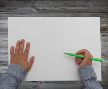Child's hands holding green marker on blank white paper on wooden surface.