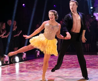 Two dancers performing energetically on stage, with one in a yellow dress and the other in a black outfit.