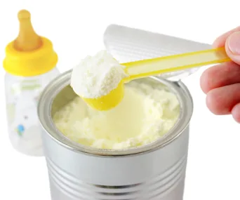 A yellow scoop of baby formula powder is held above an open can, with a baby bottle in the background.