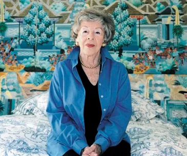 Elderly woman sitting on a bed in front of a vibrant, detailed mural, wearing a blue blouse and pearl necklace.