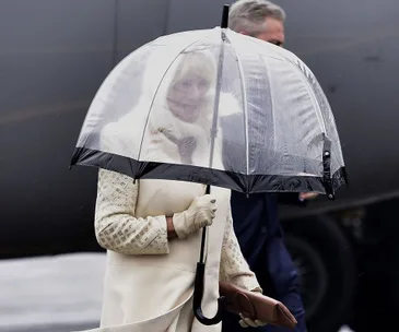 A person in a light-colored coat and gloves holds a clear umbrella, standing near a large vehicle on a rainy day.
