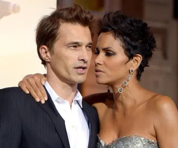 A woman in an elegant dress embraces a man in a suit on a red carpet with intimate expressions.