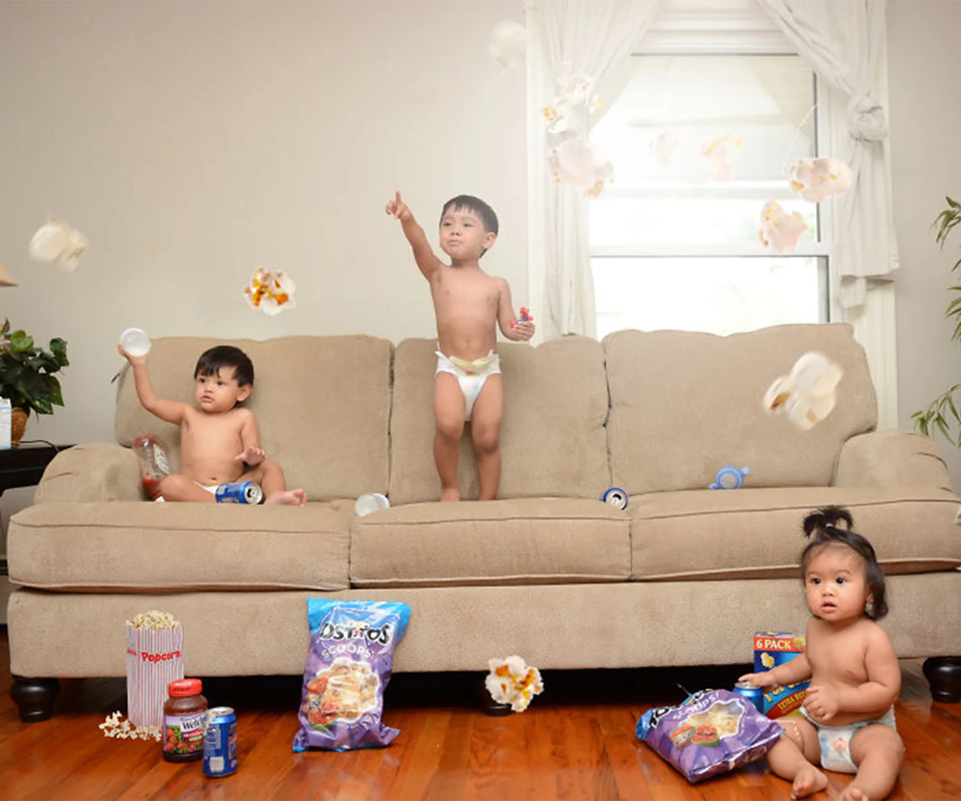 Three toddlers in diapers, tossing popcorn in a living room; snacks scattered around.