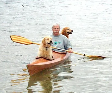 Man kayaking with two golden retrievers on a calm lake.