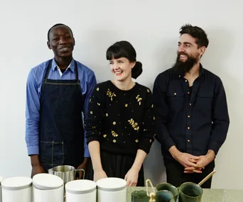 Three baristas standing behind a counter, smiling, in a coffee shop setting.