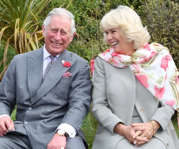 Two smiling individuals sitting outdoors, dressed in formal attire, with green foliage in the background.