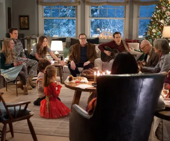 Family gathered in a cozy living room at Christmas, with a decorated tree, a guitar, and snowy scenery outside the window.