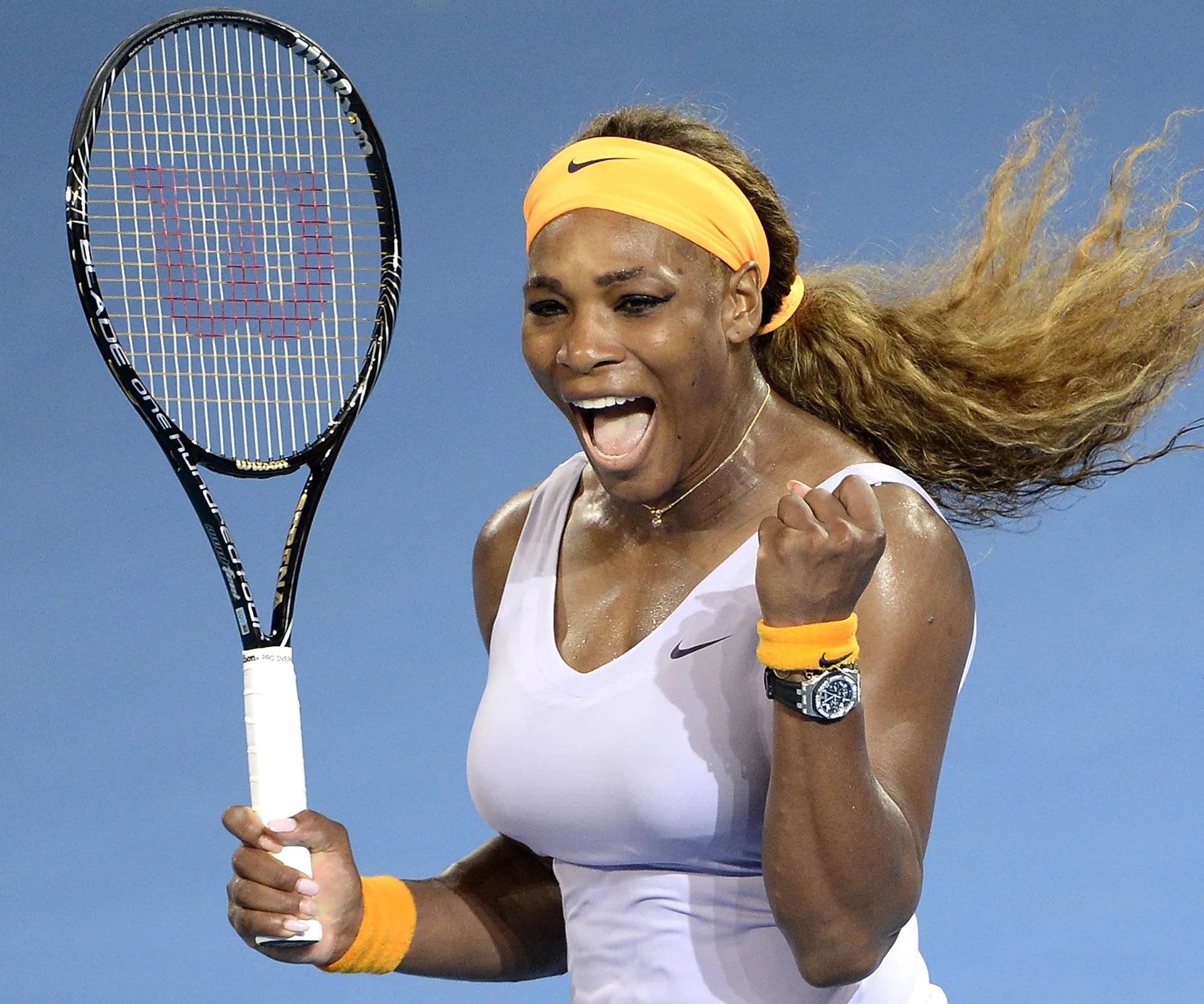 Serena Williams celebrates on court, holding a tennis racket, wearing an orange headband and wristbands.