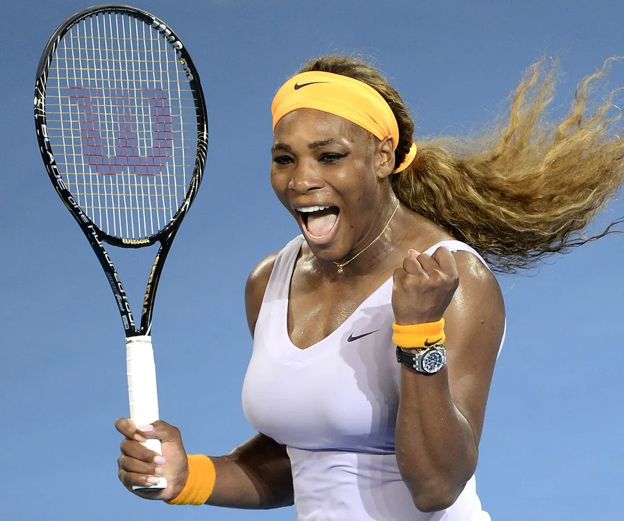 Serena Williams celebrates on court, holding a tennis racket, wearing an orange headband and wristbands.