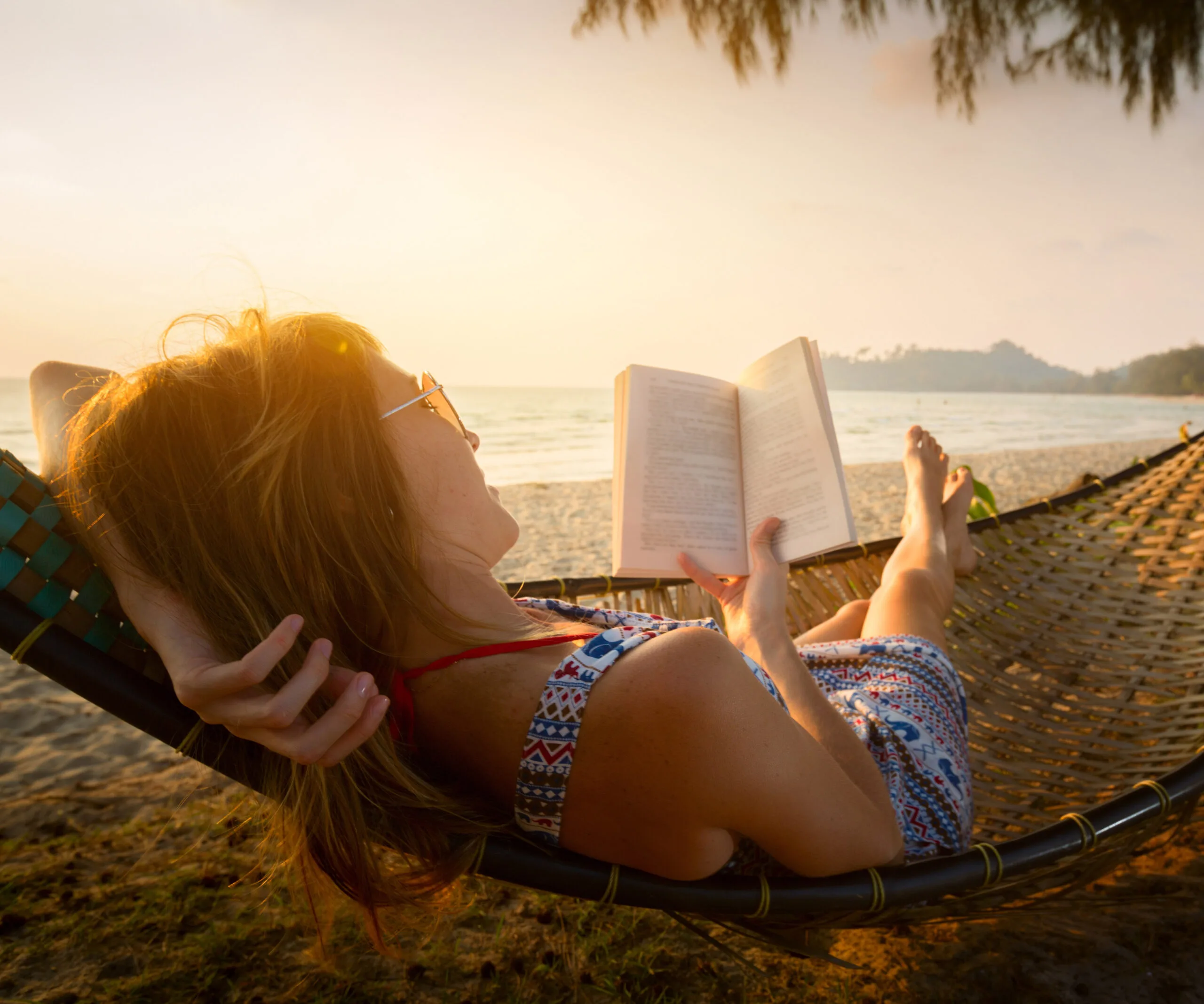 Woman reading on a hammock at the beach during sunset, dressed in a patterned summer dress.