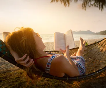 Woman reading on a hammock at the beach during sunset, dressed in a patterned summer dress.