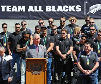 Prince Charles speaking at a podium with the New Zealand All Blacks rugby team standing behind him.