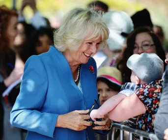 A woman in a blue outfit smiles warmly at a baby over a barrier, surrounded by a crowd in the background.