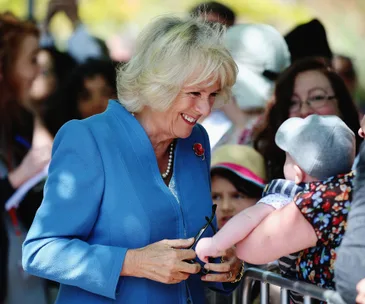 A woman in a blue outfit smiles warmly at a baby over a barrier, surrounded by a crowd in the background.
